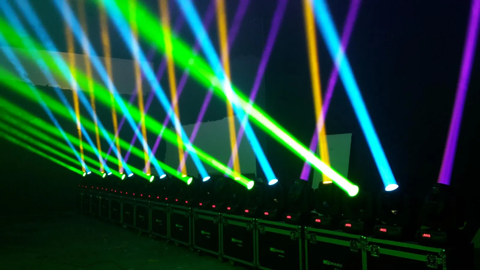 Fila de cabezas móviles encendidas sobre flight cases, con haces verdes, azules, amarillos y morados